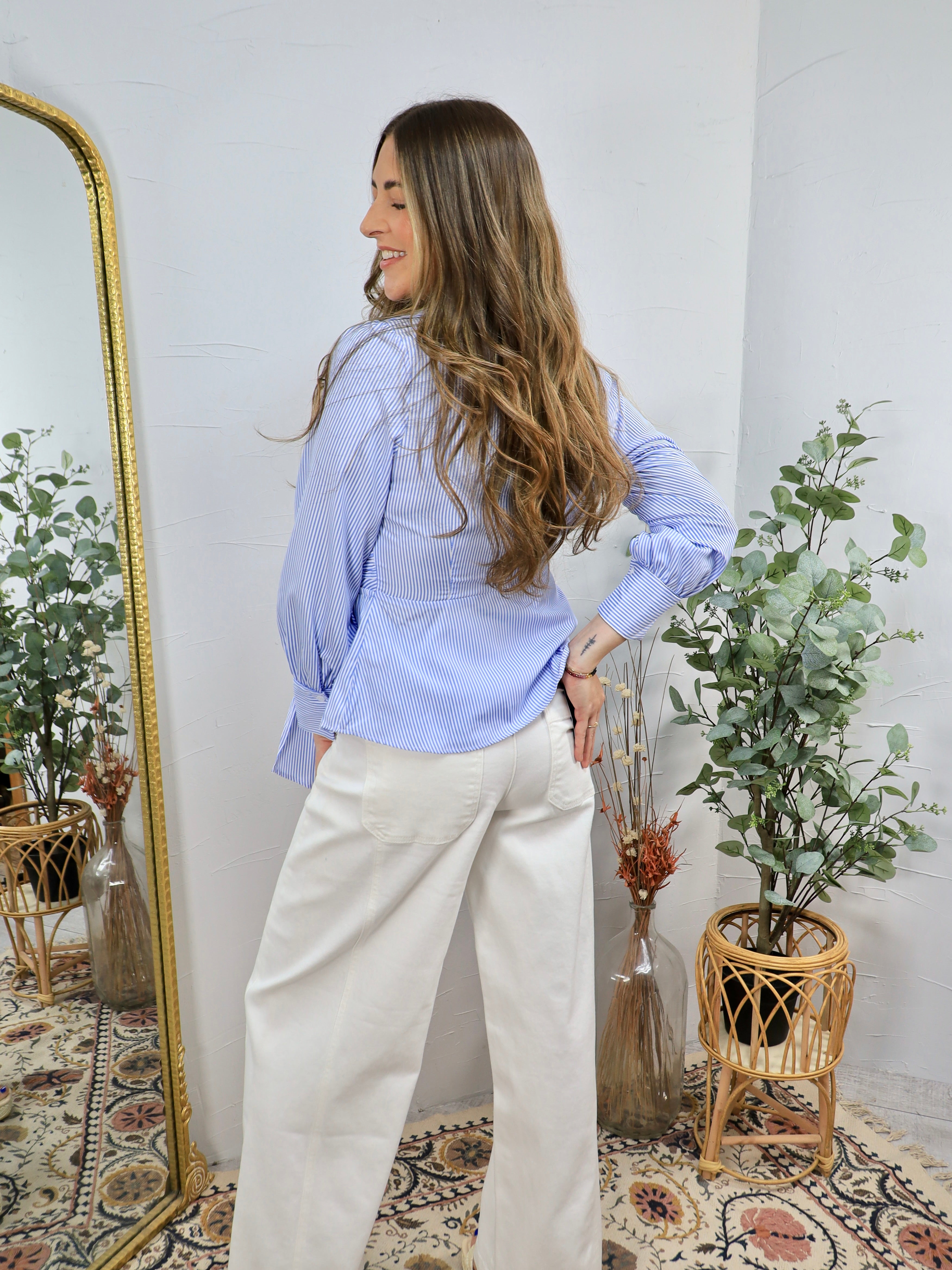 Woman wearing blue and white striped wrap blouse with white wide-leg pants, styled with long wavy hair in boho-chic setting.