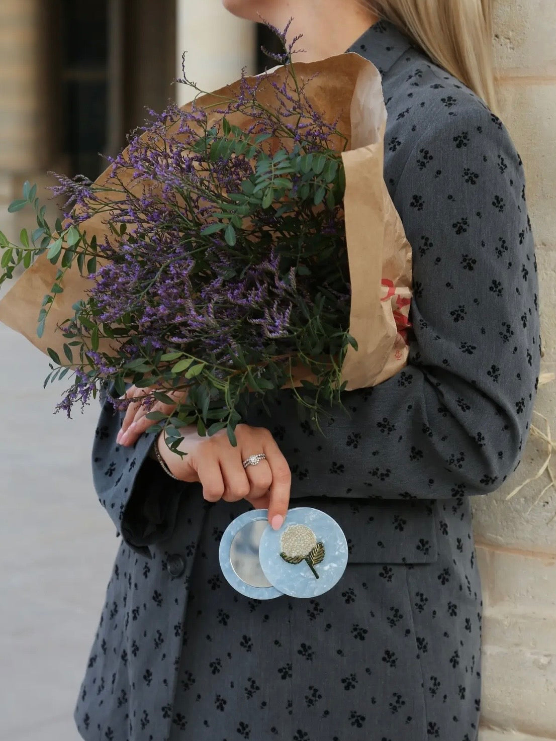 Light blue marbled compact mirror with floral brooch detail, held by woman in grey floral blazer.
