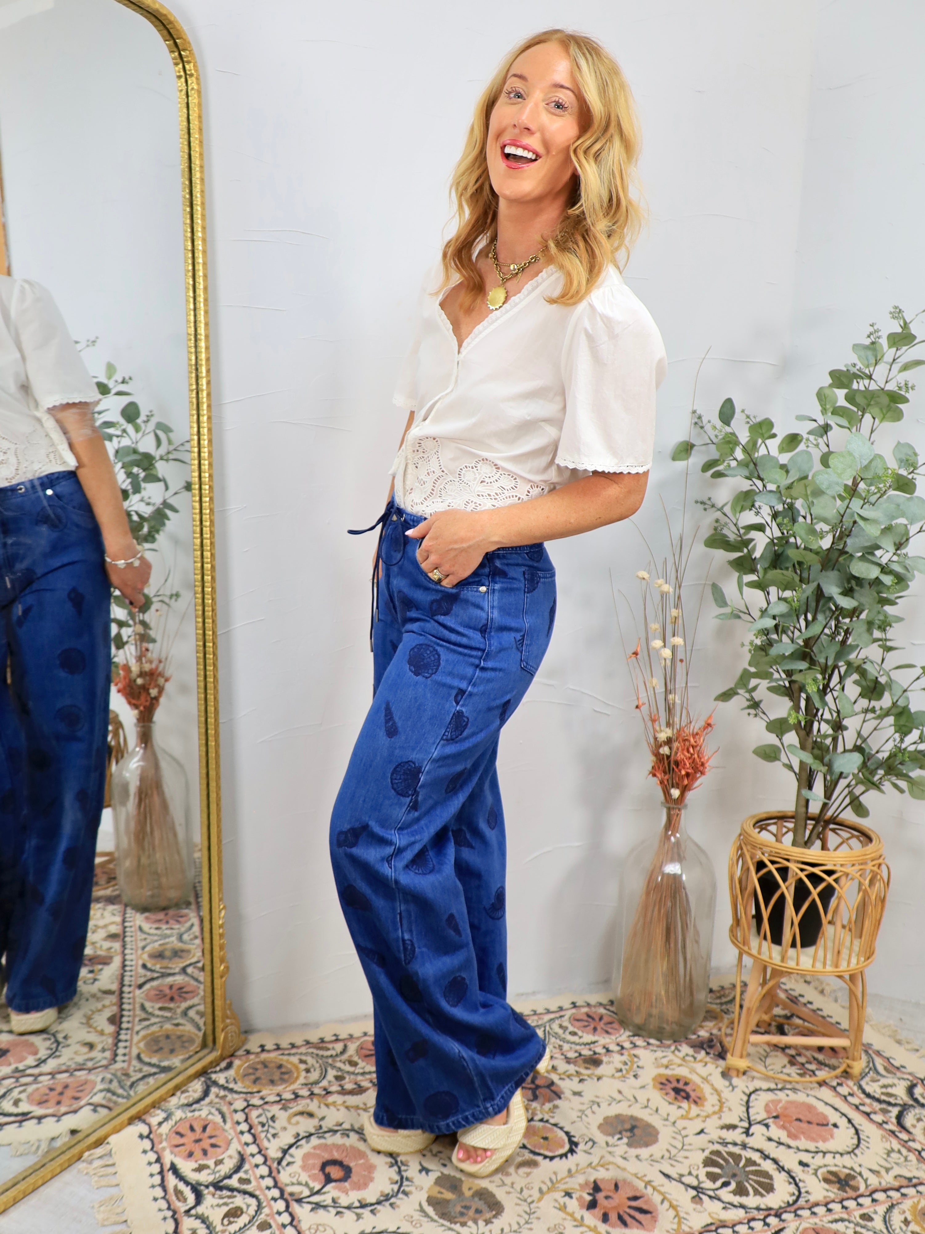 Woman wearing white cropped button-up shirt with blue wide-leg denim pants featuring shell print details, styled with layered