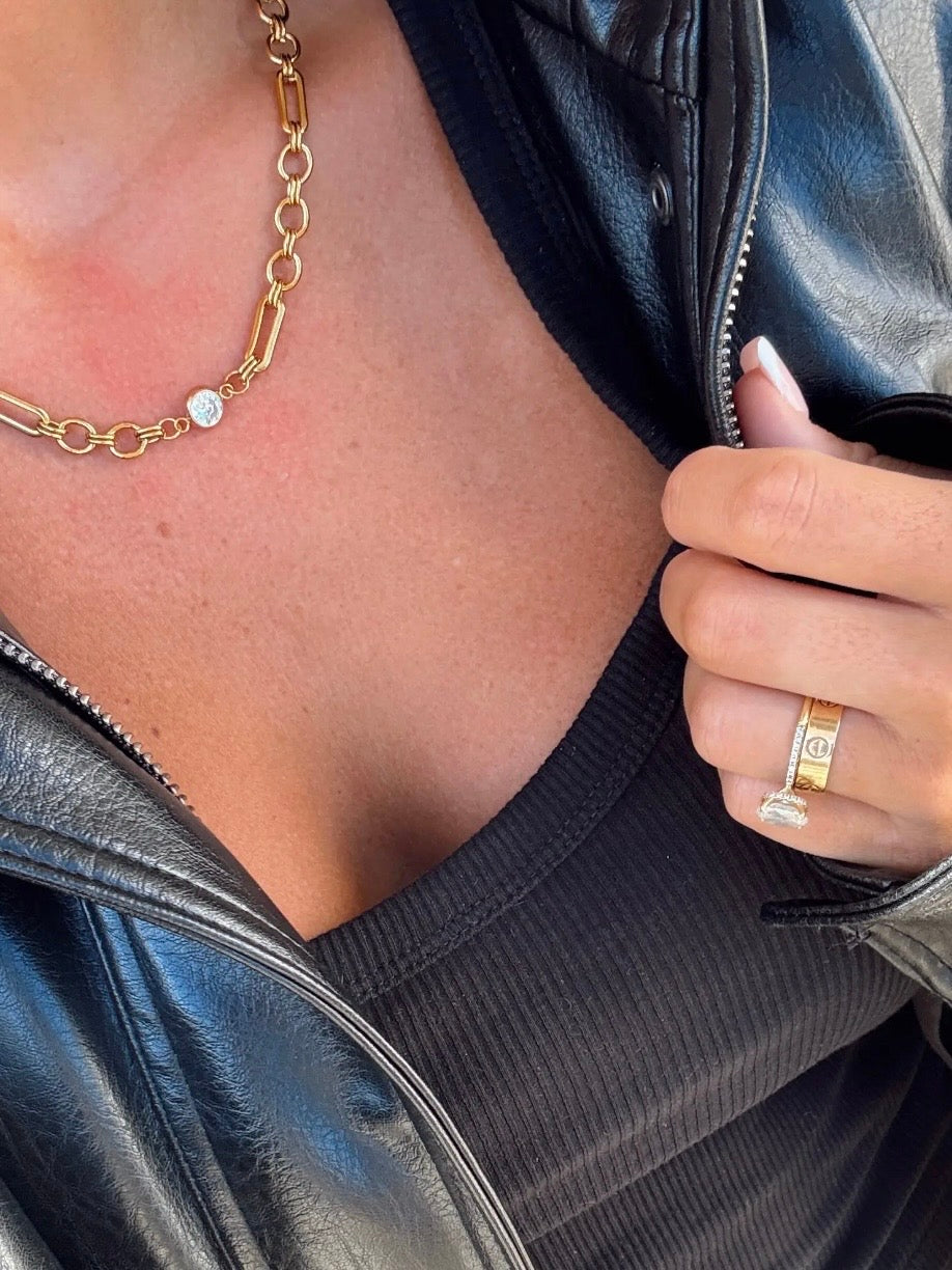 Gold mixed chain-link necklace with single bezel-set diamond pendant, worn over black ribbed top with leather jacket.