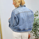 Woman wearing oversized light blue denim jacket with white pants, shown from behind in minimal home setting with plants.