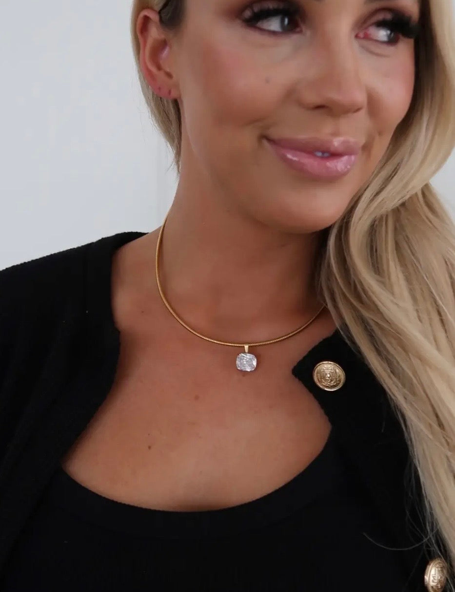 Gold snake chain necklace with cushion-cut crystal pendant, styled on model wearing black blazer.