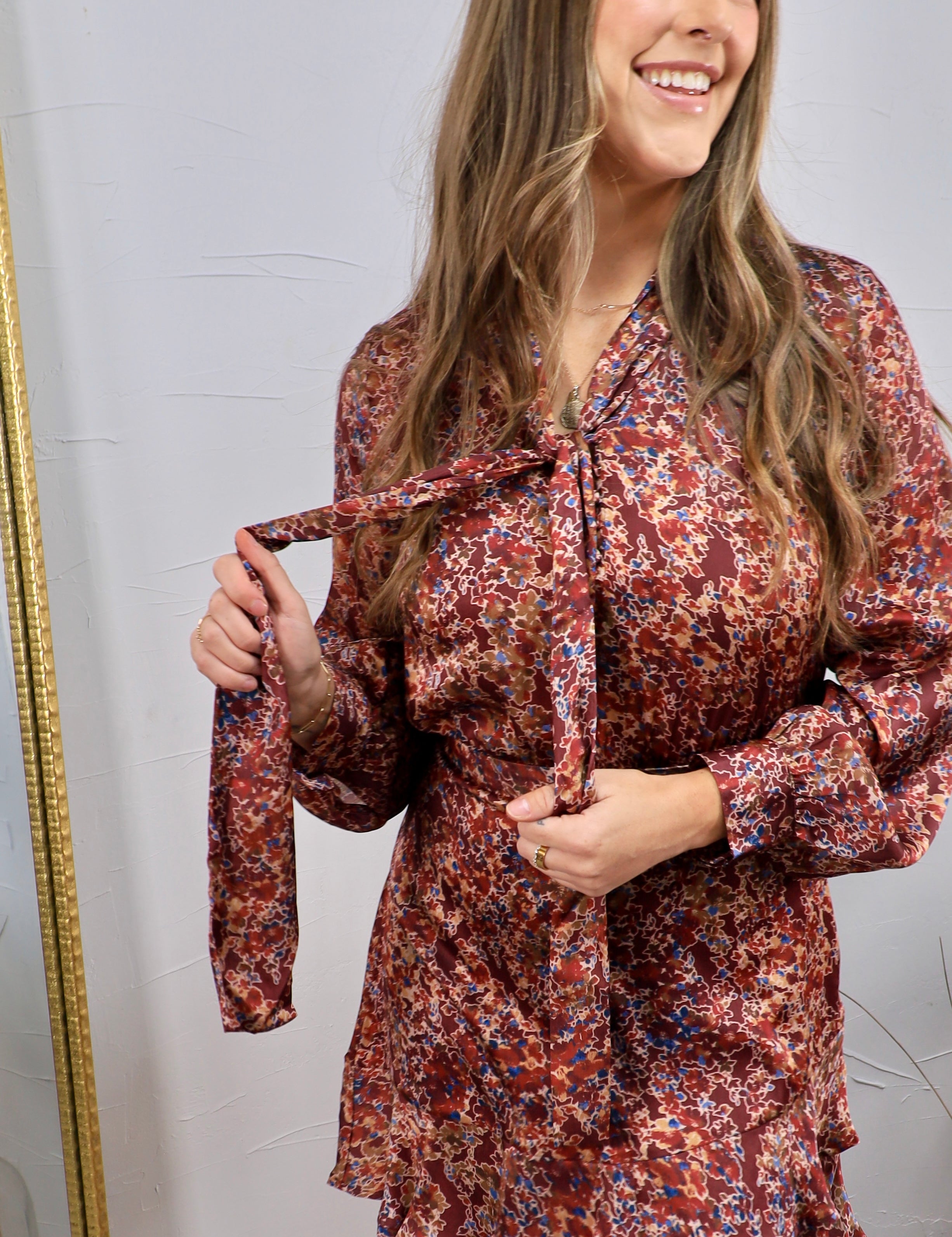 Burgundy floral satin mini dress with long sleeves, tie neck detail, and ruffle hem, shown on model.