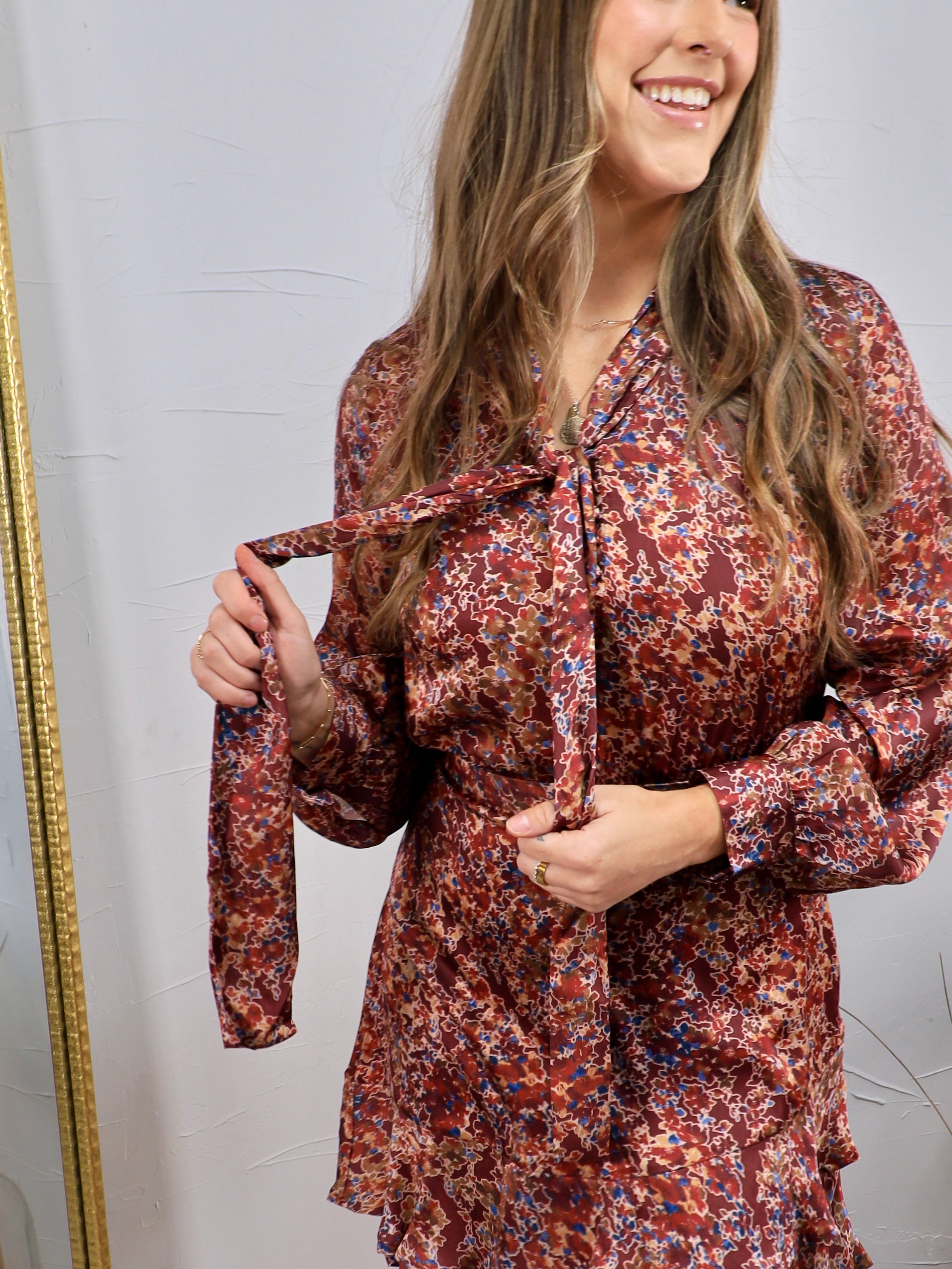 Burgundy floral satin mini dress with long sleeves, tie neck detail, and ruffle hem, shown on model.