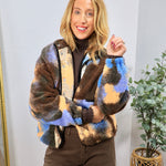 Multicolor sherpa bomber jacket in blue, brown, and tan camo print, styled over dark turtleneck and brown wide-leg pants.
