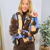 Multicolor sherpa bomber jacket in blue, brown, and tan camo print, styled over dark turtleneck and brown wide-leg pants.