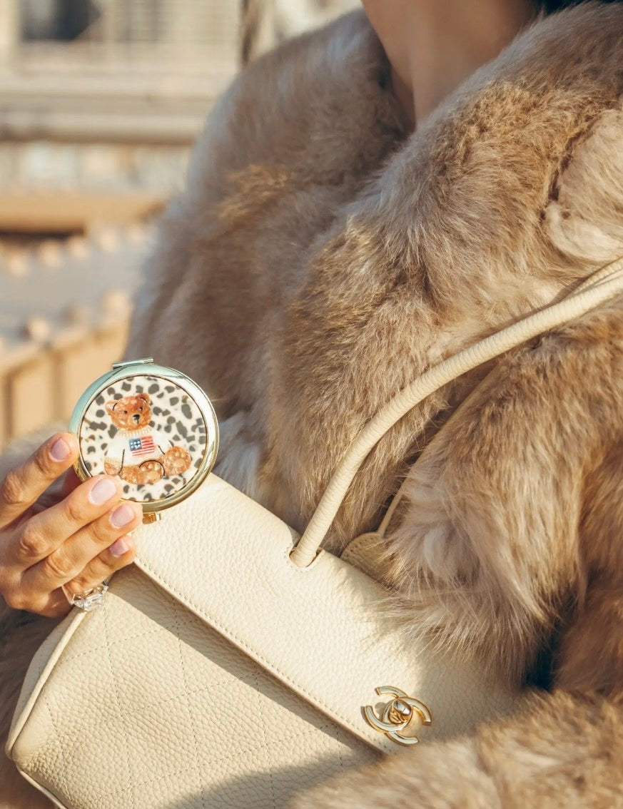 Round white and gold teddy bear compact held in hand alongside cream Chanel bag and fur coat