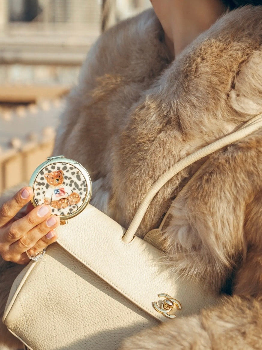 Round white and gold teddy bear compact held in hand alongside cream Chanel bag and fur coat