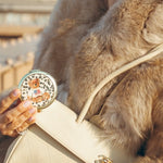Round white and gold teddy bear compact held in hand alongside cream Chanel bag and fur coat