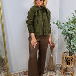 Olive green suede button-front jacket with patch pockets, styled over brown wide-leg trousers and ankle boots.