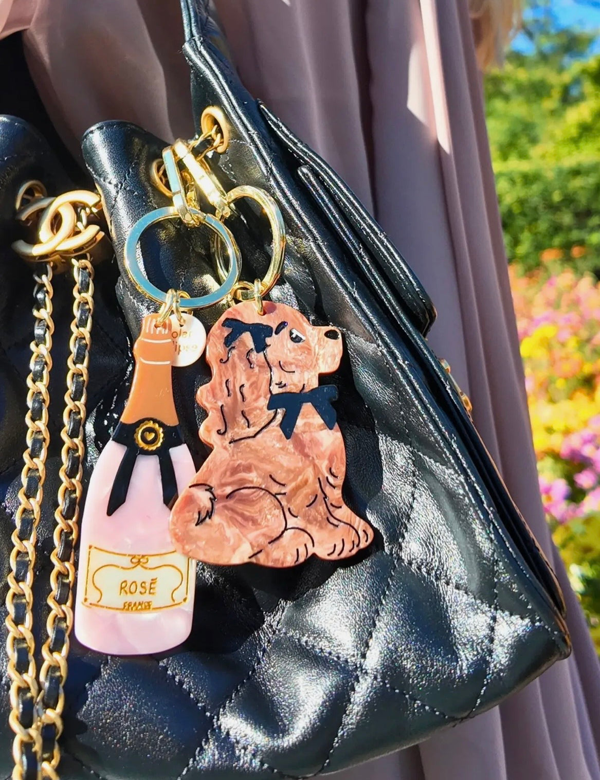 Two acrylic keychains featuring a rosé bottle and Cocker Spaniel charm on a black quilted handbag