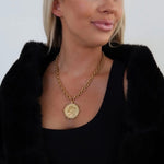 Gold coin pendant necklace on chunky link chain, worn with black top and faux fur jacket.
