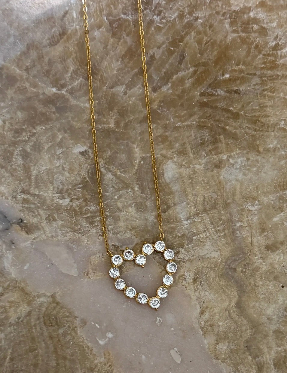 Gold chain necklace with open heart pendant formed by round sparkling clear crystal stones on marble surface.