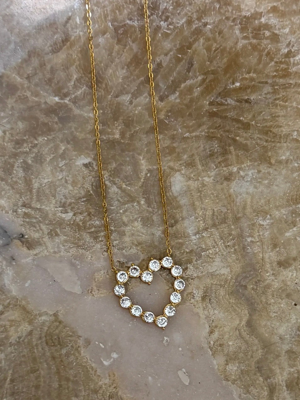 Gold chain necklace with open heart pendant formed by round sparkling clear crystal stones on marble surface.