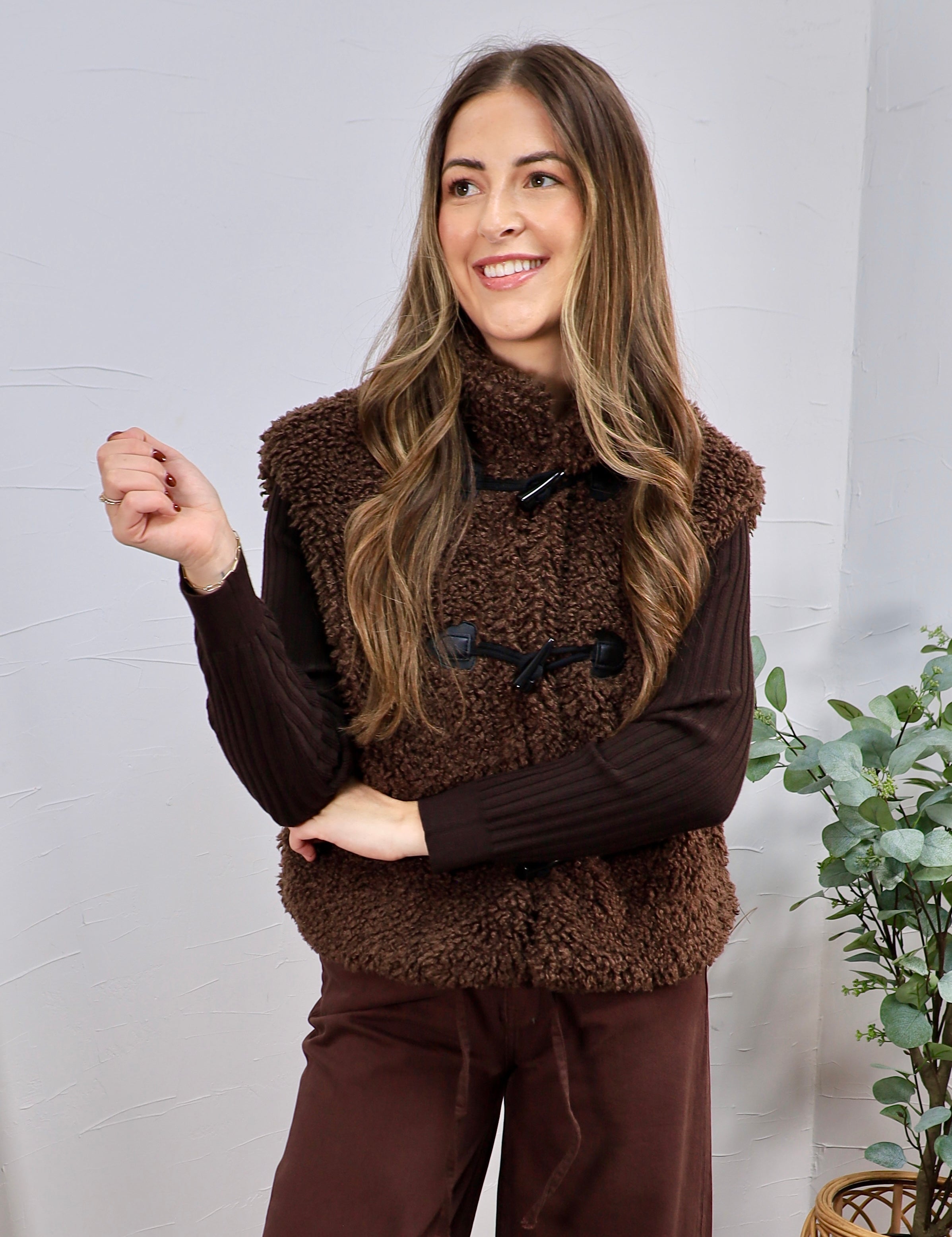 Brown teddy fleece vest with black toggle closures, styled over a dark brown ribbed long-sleeve sweater.