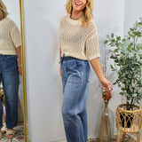 Woman wearing beige crochet knit sweater with short sleeves paired with high-waisted blue jeans, standing by mirror.