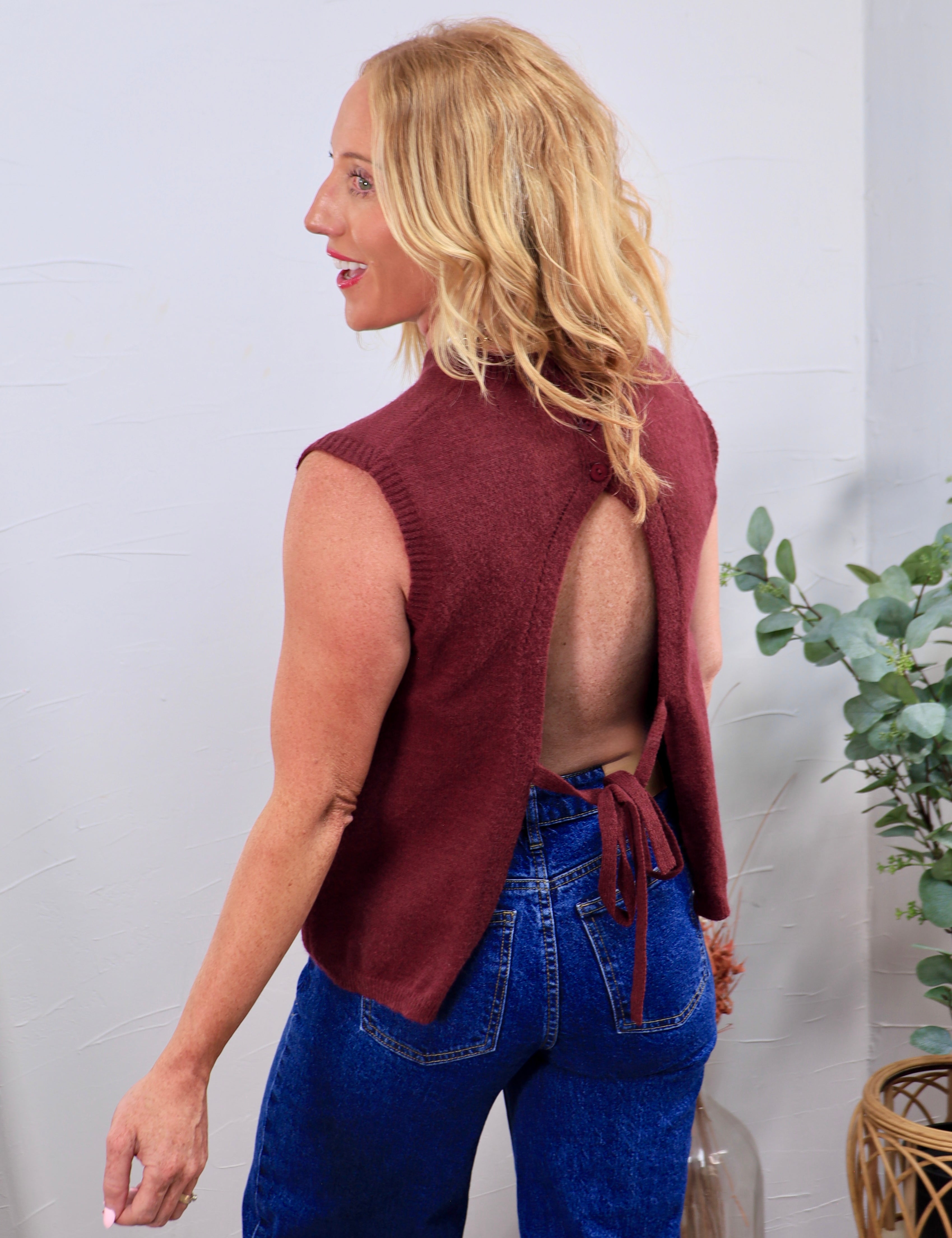 Burgundy open-back knit vest with tie closure, styled over blue high-waisted jeans, shown from behind.