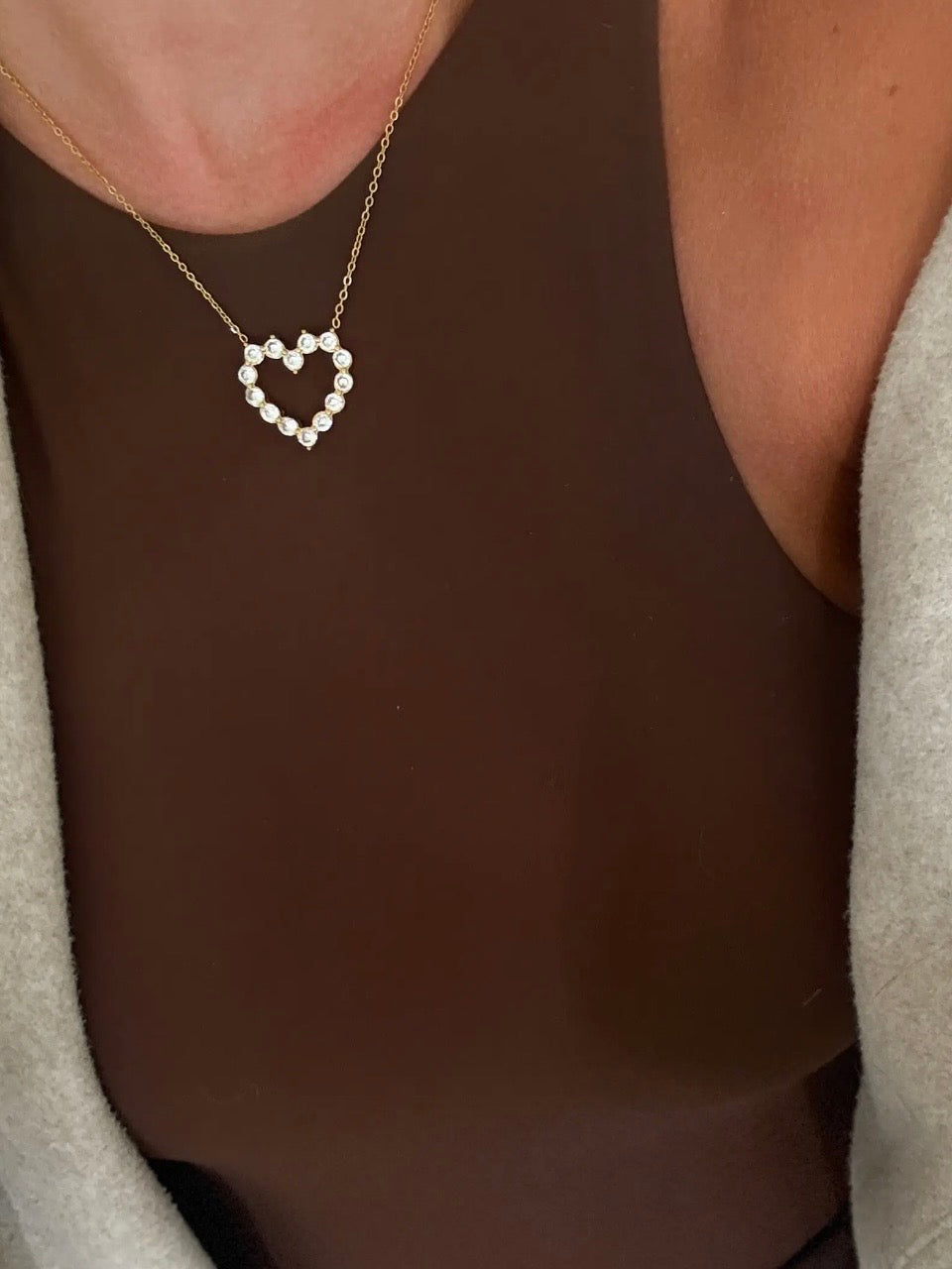 Gold chain necklace with open heart pendant set with round diamond stones, worn over brown top.