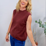Woman wearing a burgundy sleeveless knit sweater vest with blue jeans, smiling against a light gray wall
