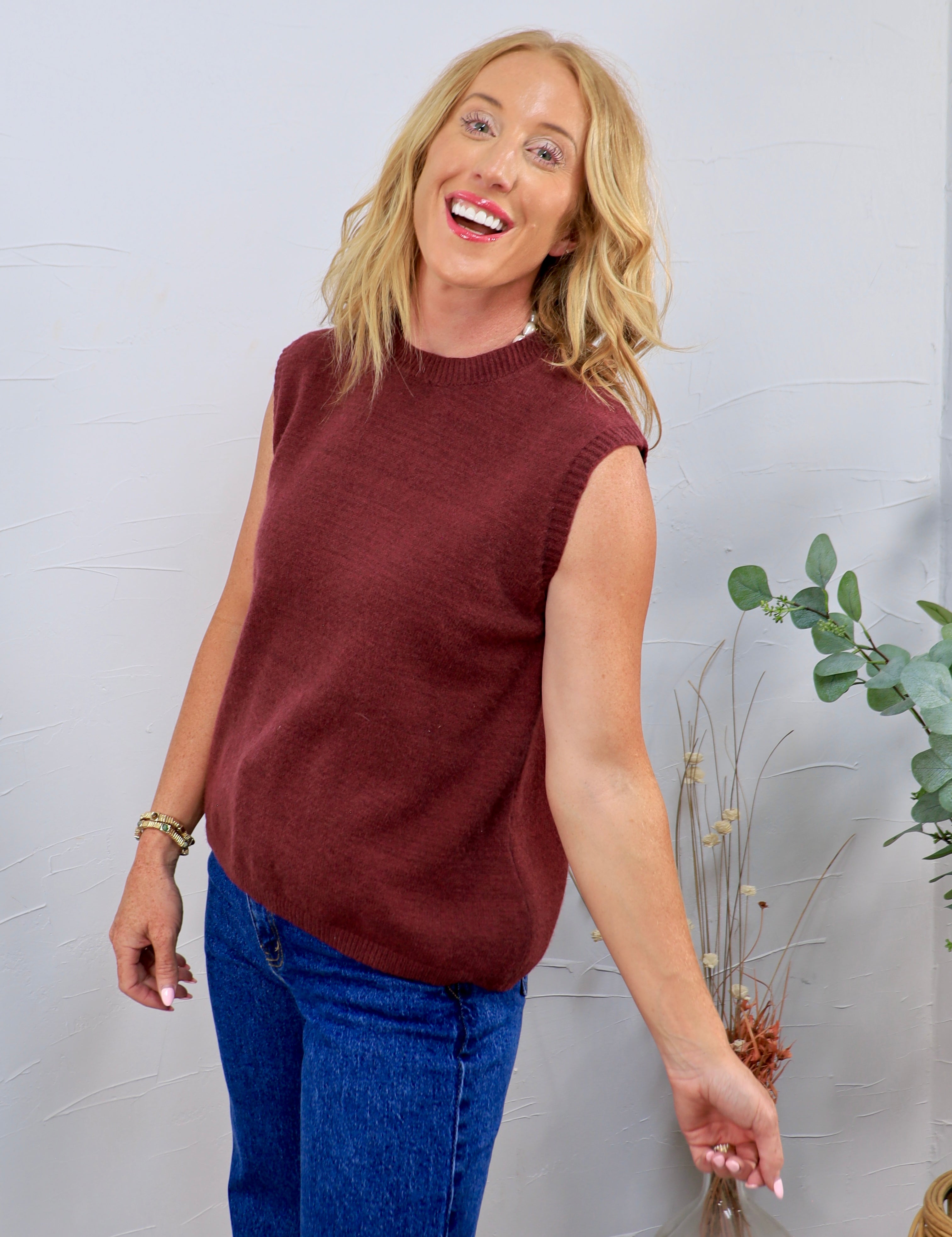 Woman wearing a burgundy sleeveless knit sweater vest with blue jeans, smiling against a light gray wall