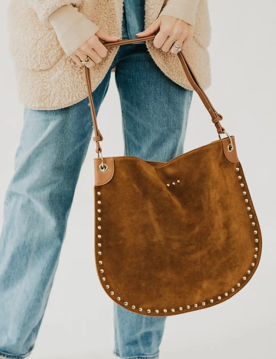 Tan suede hobo tote bag with gold stud trim, held by model wearing blue jeans and cream sherpa jacket.