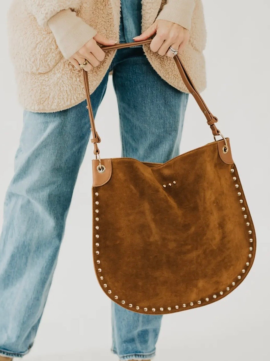 Tan suede hobo tote bag with gold stud trim, held by model wearing blue jeans and cream sherpa jacket.