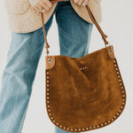 Tan suede hobo tote bag with gold stud trim, held by model wearing blue jeans and cream sherpa jacket.
