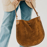 Tan suede hobo tote bag with gold stud trim, held by model wearing blue jeans and cream sherpa jacket.