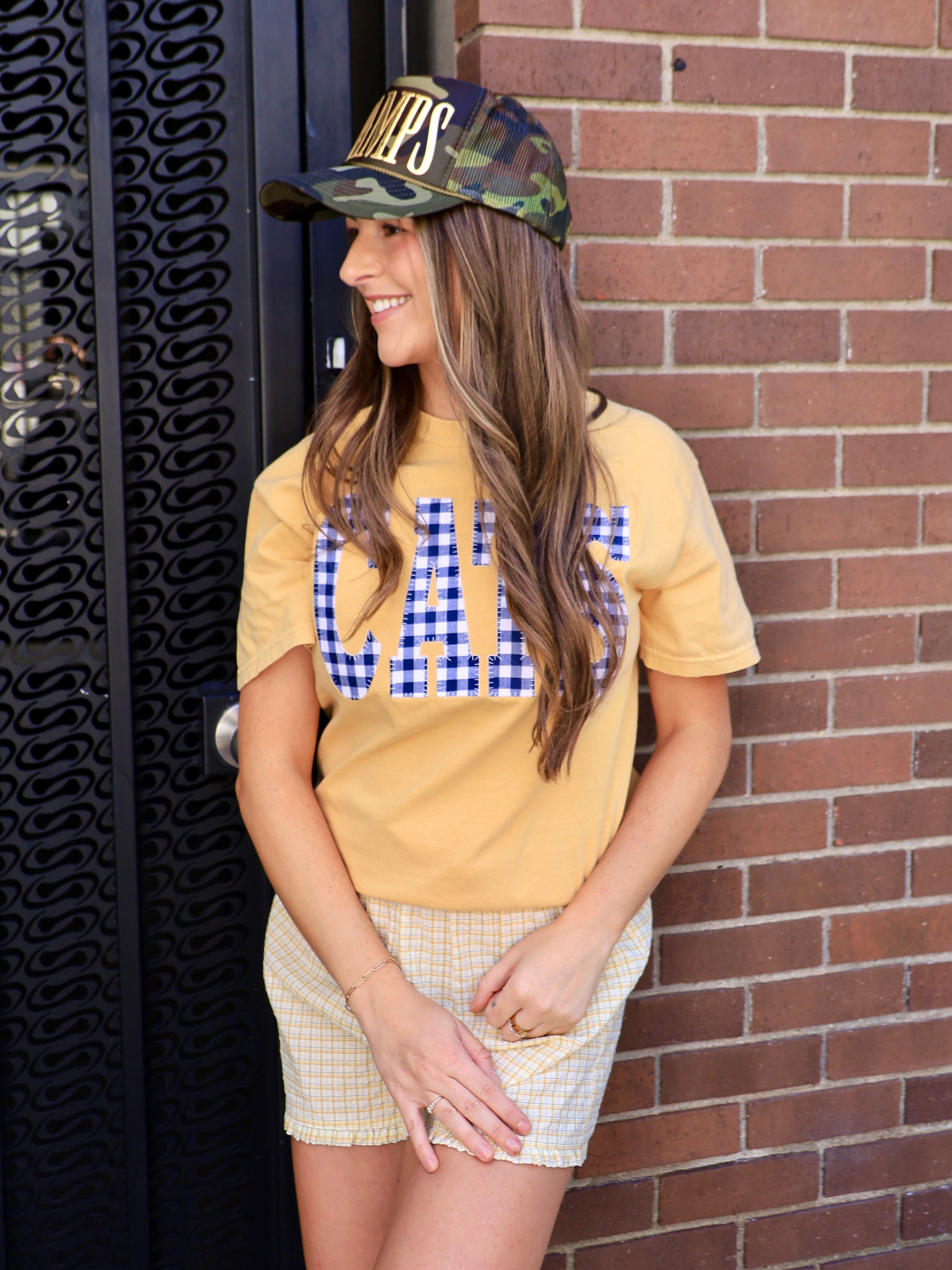 Camouflage trucker hat with gold "CHAMPS" text, styled with yellow graphic tee and plaid shorts.