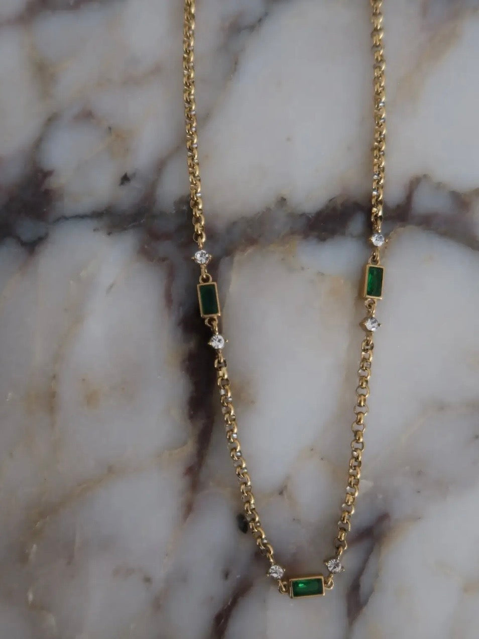 Gold chain necklace featuring alternating emerald green baguette stones and clear crystal accents on marble surface.