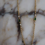 Gold chain necklace featuring alternating emerald green baguette stones and clear crystal accents on marble surface.