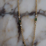 Gold chain necklace featuring alternating emerald green baguette stones and clear crystal accents on marble surface.