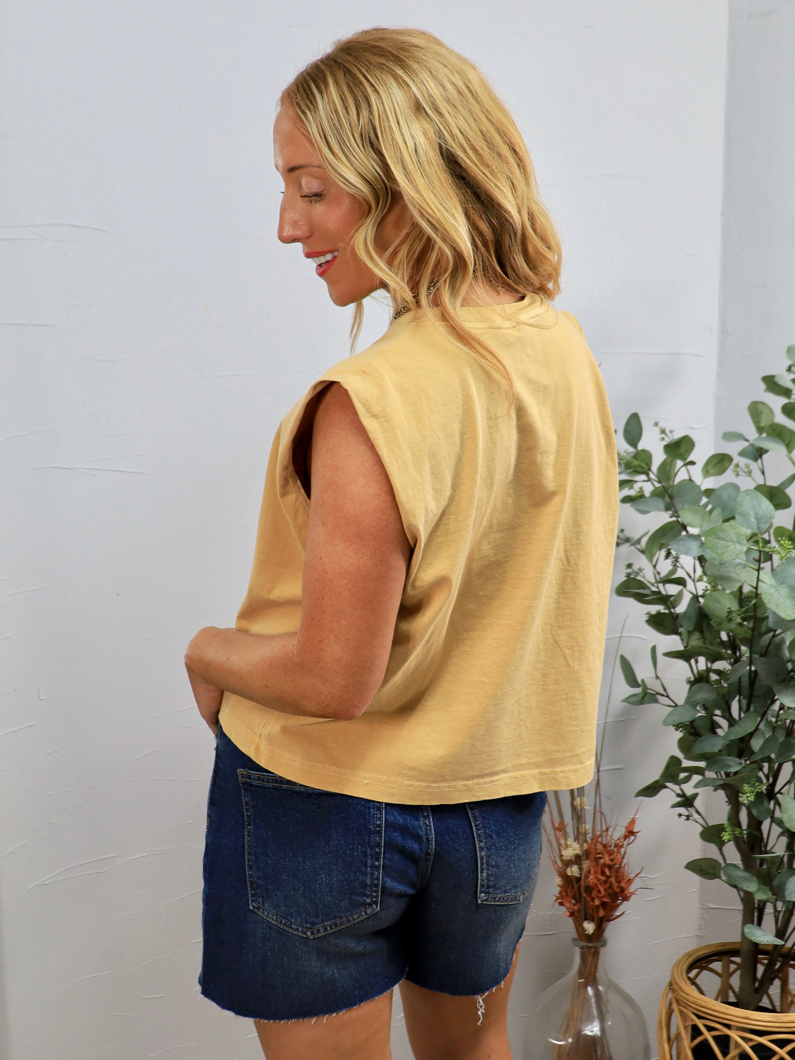 Woman wearing yellow sleeveless muscle tee with blue denim shorts, shown from behind in casual styling setup.