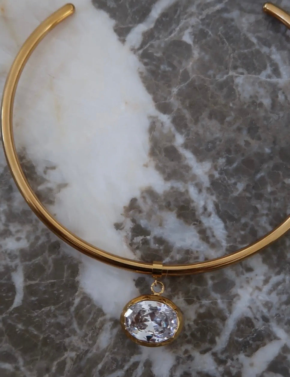 Gold rigid choker necklace with oval crystal pendant displayed on white and grey marble surface.
