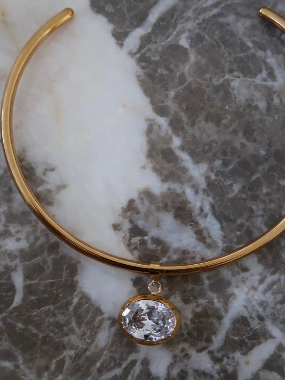 Gold rigid choker necklace with oval crystal pendant displayed on white and grey marble surface.