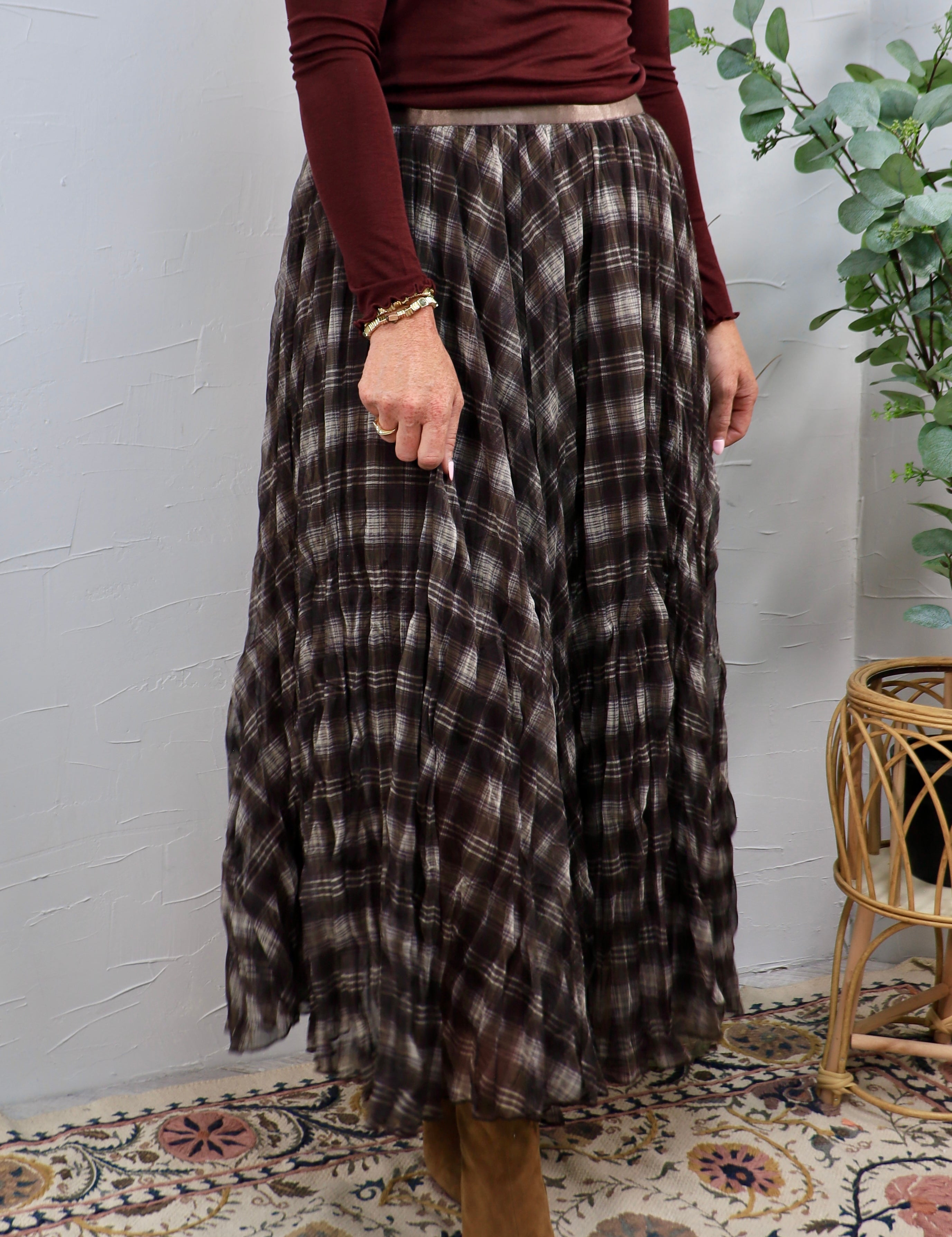 Woman wearing a dark brown plaid pleated maxi skirt with a burgundy long-sleeve top and gold bracelet