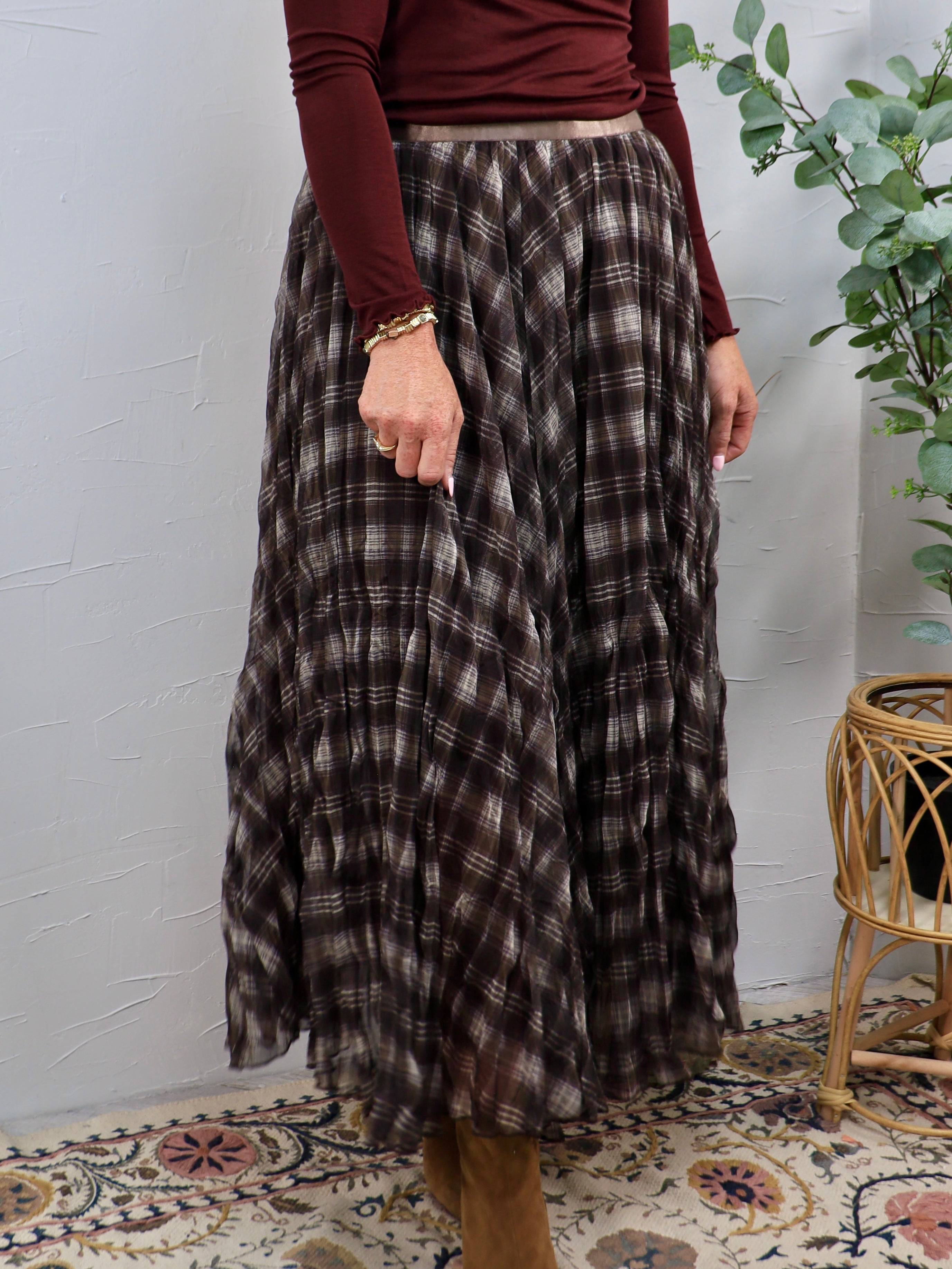 Woman wearing a dark brown plaid pleated maxi skirt with a burgundy long-sleeve top and gold bracelet