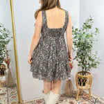 Woman wearing black floral print sleeveless mini dress with square neckline and white cowboy boots, shown from behind.