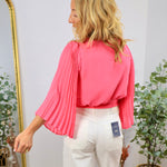 Coral pink wrap blouse with pleated flutter sleeves, styled with white wide-leg jeans, back view.
