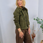 Olive green suede button-front jacket with patch pockets, styled over brown wide-leg pants with gold bracelets.