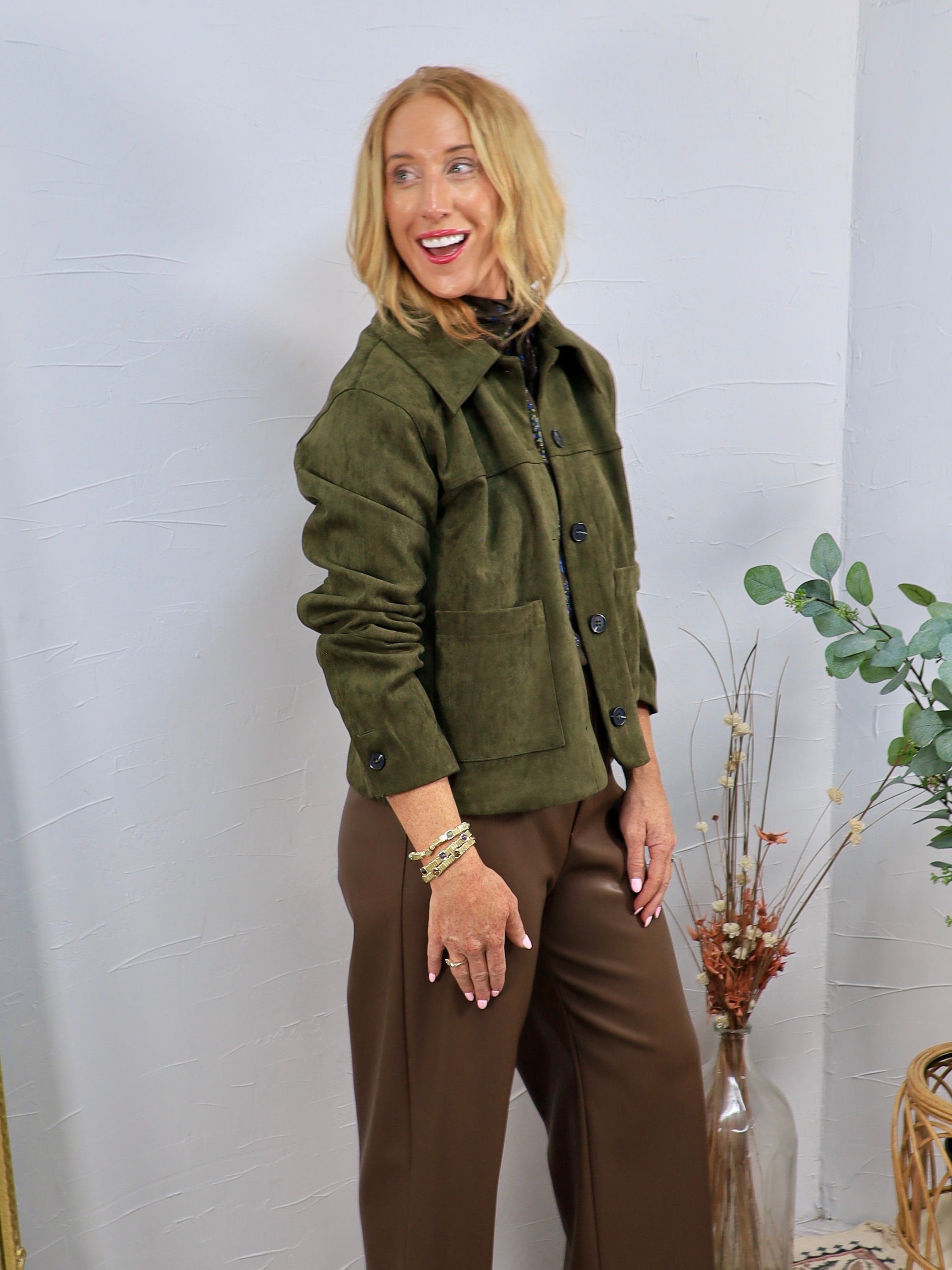 Olive green suede button-front jacket with patch pockets, styled over brown wide-leg pants with gold bracelets.