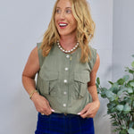 Sage green sleeveless utility top with white buttons and chest pockets, styled with dark blue jeans.