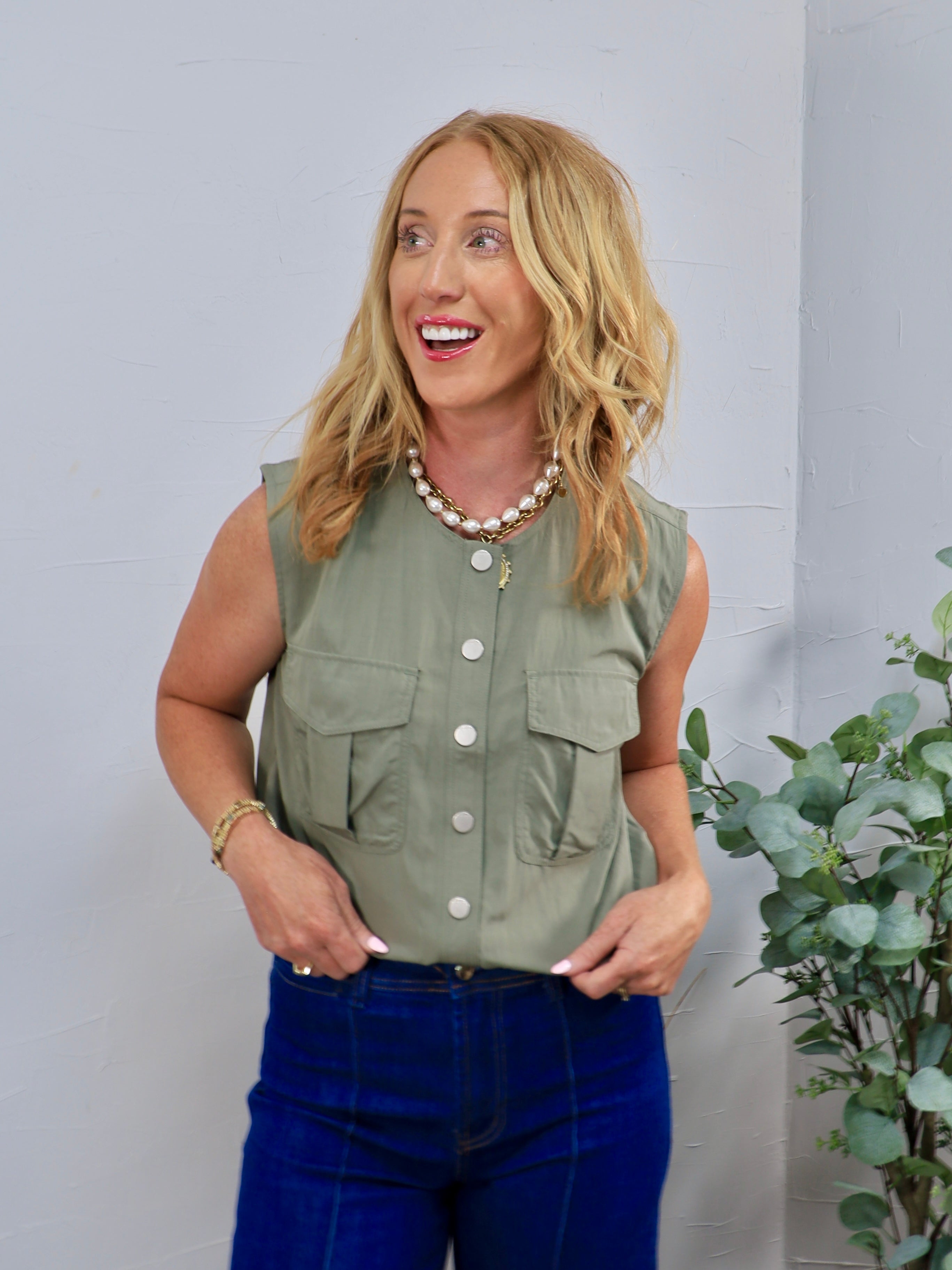 Sage green sleeveless utility top with white buttons and chest pockets, styled with dark blue jeans.