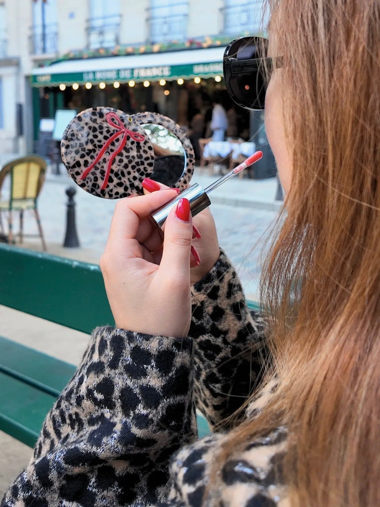 Woman holding a leopard print compact mirror with red bow and lip gloss in Paris