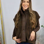 Brown sherpa fleece vest with snap buttons, styled over a dark brown long-sleeve top and blue jeans.