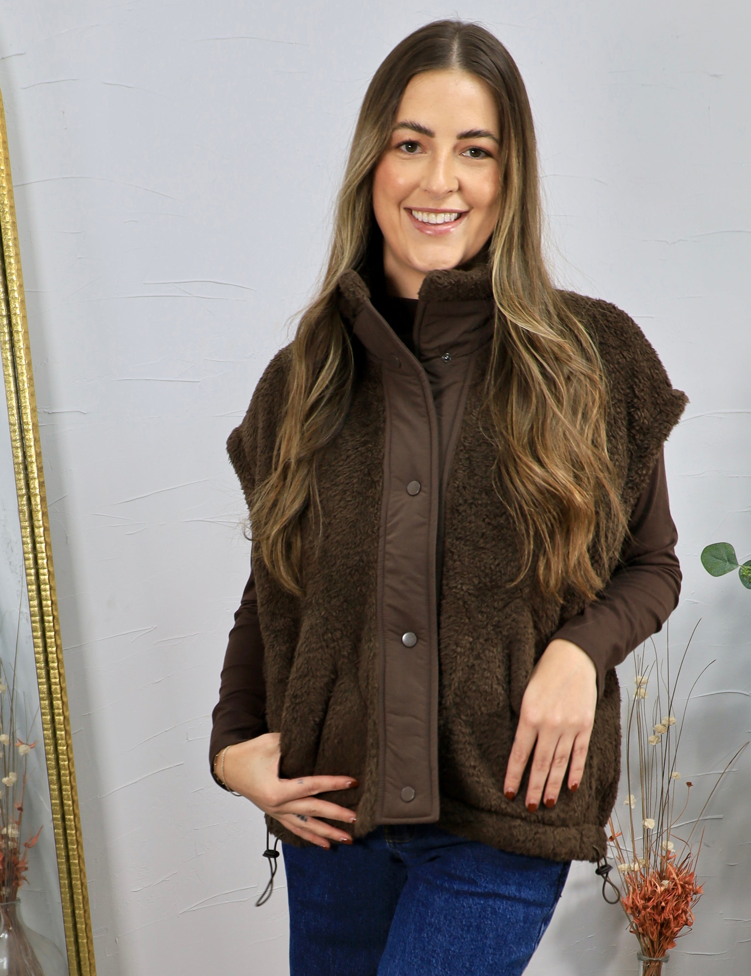 Brown sherpa fleece vest with snap buttons, styled over a dark brown long-sleeve top and blue jeans.