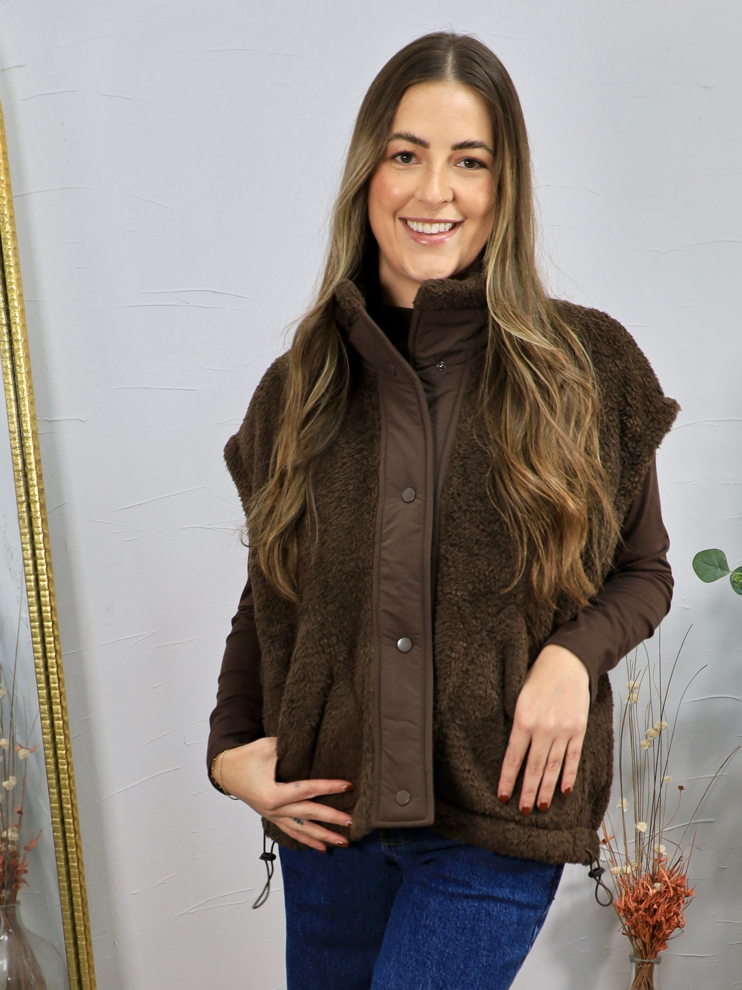 Brown sherpa fleece vest with snap buttons, styled over a dark brown long-sleeve top and blue jeans.