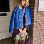Woman wearing cropped sherpa-lined denim jacket over brown maxi dress holding camo clutch outdoors