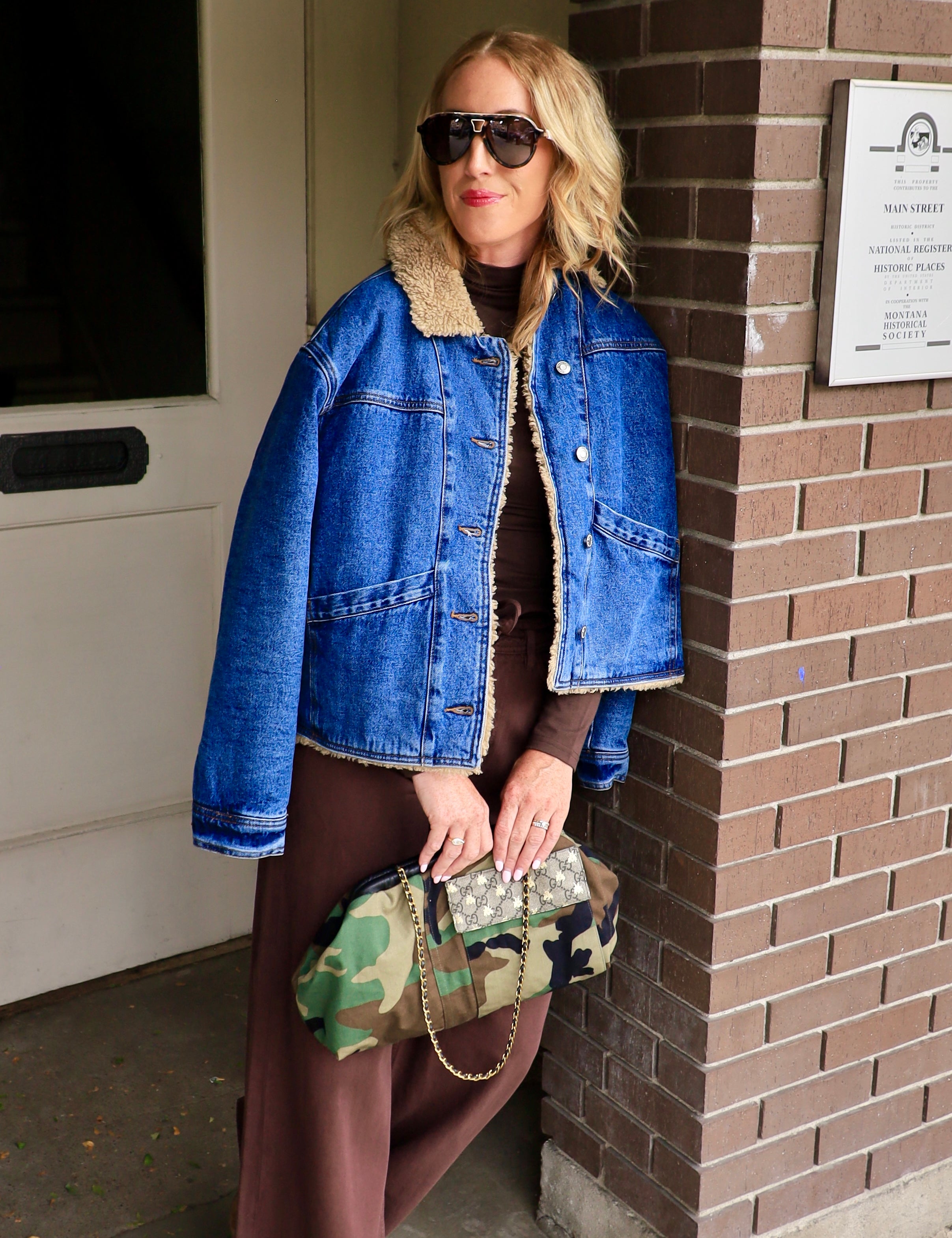 Woman wearing cropped sherpa-lined denim jacket over brown maxi dress holding camo clutch outdoors