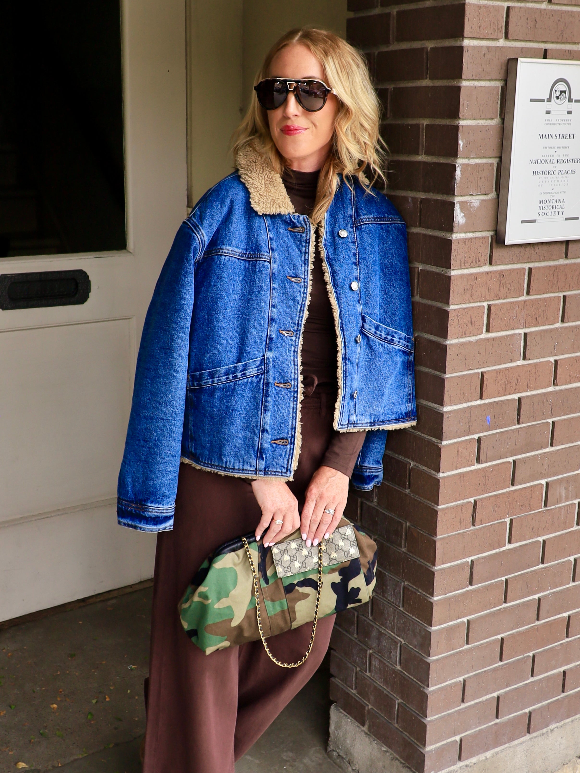 Woman wearing cropped sherpa-lined denim jacket over brown maxi dress holding camo clutch outdoors